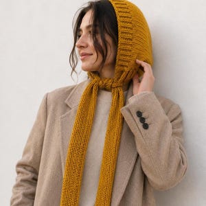 May include: A mustard yellow knitted hood with long ties that knot under the chin. The hood features a ribbed texture along the front edge and is crafted from a thick yarn, providing full head coverage with two long, dangling knitted scarf-like ends.