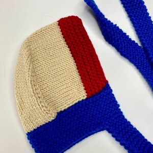 May include: A hand-knitted bonnet featuring a color-blocked design with a large cream-colored section, a vertical red stripe, and a vibrant blue base. The bonnet includes two long, matching blue knitted ties extending from the bottom edge.