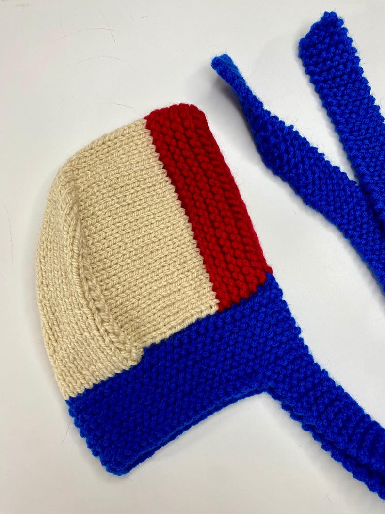May include: A hand-knitted bonnet featuring a color-blocked design with a large cream-colored section, a vertical red stripe, and a vibrant blue base. The bonnet includes two long, matching blue knitted ties extending from the bottom edge.