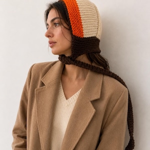 May include: A knitted bonnet featuring a cream-colored crown, a vibrant orange band, and a dark brown base with long, matching brown ties. The bonnet is worn with a beige V-neck sweater layered under a tan wool-blend blazer with notched lapels.