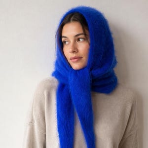 May include: A vibrant royal blue headscarf crafted from a soft, fuzzy, mohair-like knit material. The scarf is draped over the head and tied loosely under the chin, featuring long, pointed ends that hang down in front of the chest.