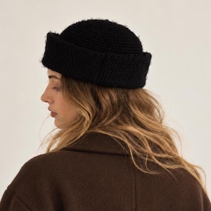 May include: A solid black beanie with a thick, folded cuff. The hat features a textured, crocheted knit construction with a rounded crown that sits high on the head. The material appears to have a slightly fuzzy, soft surface texture throughout.