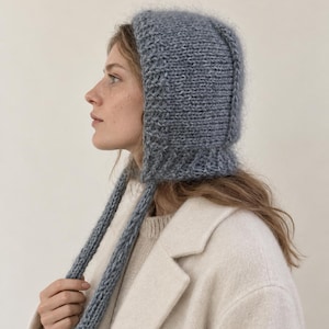 May include: A blue, chunky-knit bonnet with a fuzzy, textured surface and long, dangling knitted ties. The headwear features a ribbed border along the face opening and is crafted from a thick, soft yarn, providing full coverage for the head.