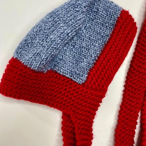 May include: A hand-knitted bonnet featuring a vibrant red base with a contrasting light blue panel on the crown. The bonnet is constructed with a textured garter stitch pattern and includes two long, thick red knitted ties attached at the sides.