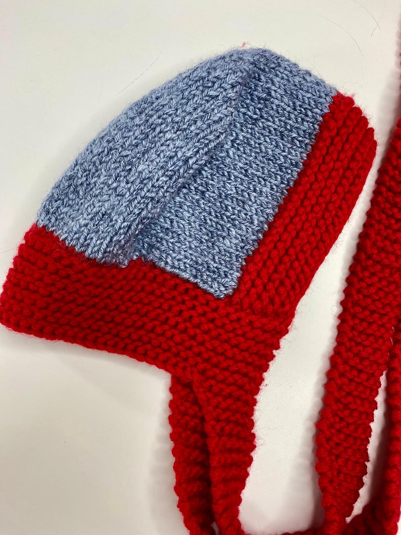 May include: A hand-knitted bonnet featuring a vibrant red base with a contrasting light blue panel on the crown. The bonnet is constructed with a textured garter stitch pattern and includes two long, thick red knitted ties attached at the sides.