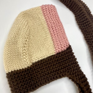 May include: A hand-knitted bonnet featuring a color-blocked design with a cream-colored main section, a vertical stripe of soft pink, and a dark brown border along the bottom edge. The bonnet includes long, matching dark brown knitted ties.