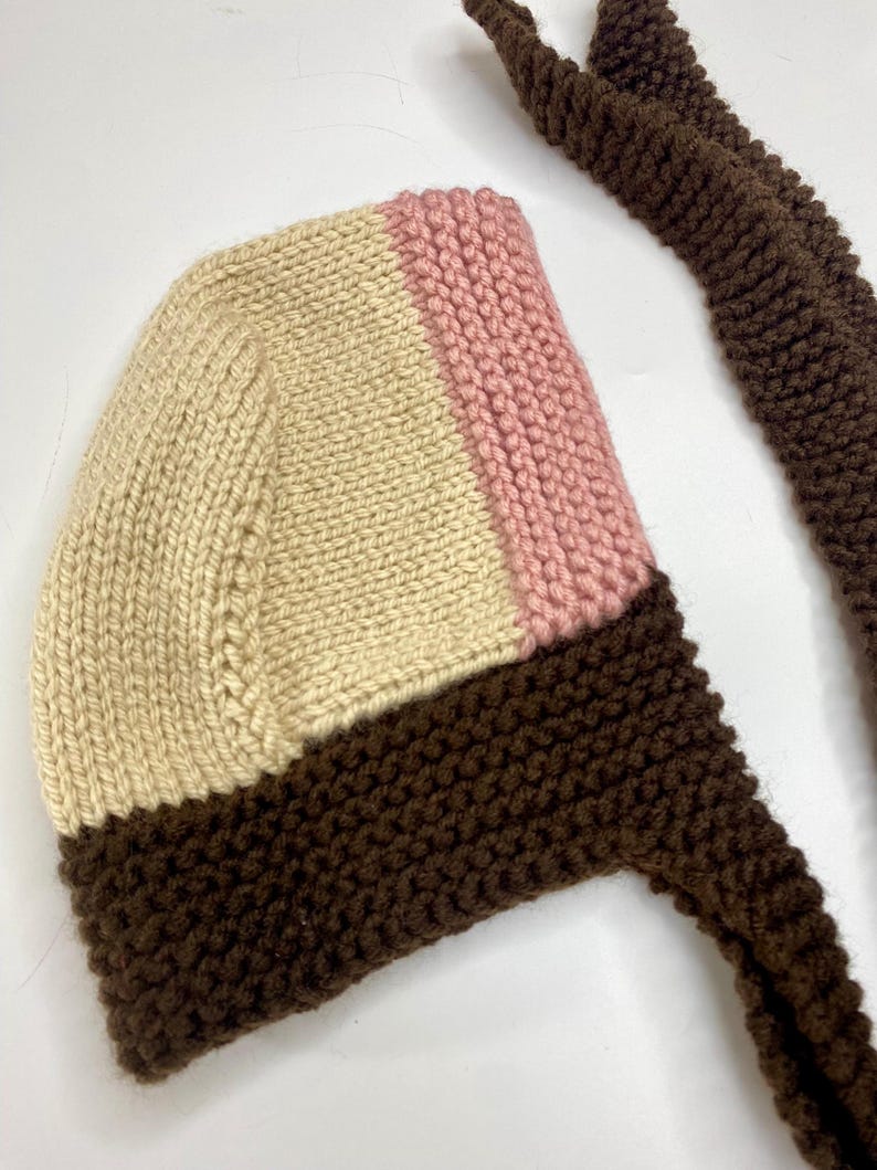 May include: A hand-knitted bonnet featuring a color-blocked design with a cream-colored main section, a vertical stripe of soft pink, and a dark brown border along the bottom edge. The bonnet includes long, matching dark brown knitted ties.