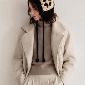 May include: A crochet bonnet featuring a black and cream granny square pattern. The headwear includes two long black braided ties that hang down from the sides, each finished with a small black pom-pom at the end of the cord.