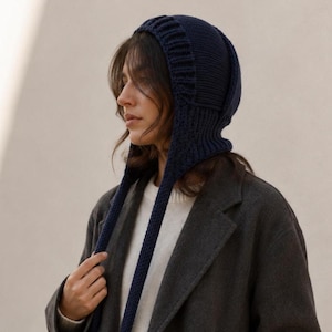 May include: A dark navy blue knitted balaclava-style bonnet featuring a ribbed texture along the front edge and two long, narrow knitted ties hanging down from the sides. The headwear is constructed with a dense, uniform stitch pattern.