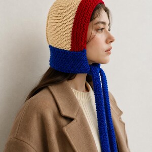 May include: A knit bonnet featuring a color-blocked design with a beige crown, a red band around the face, and a blue lower section with long, thick blue ties that hang down. The hat is constructed with a visible textured knit stitch pattern.