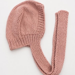 May include: A dusty rose-colored knitted bonnet featuring a rounded crown and a ribbed texture along the edges. The headwear includes a long, integrated knitted strap that extends from the base, showcasing a consistent stockinette stitch pattern.