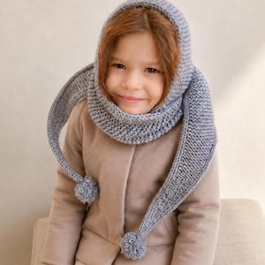 Könnte beinhalten: Eine graue Strickkapuze mit strukturiertem Strickmuster, die sich wie ein Schal um den Hals legt. Die Kapuze hat zwei lange, spitz zulaufende Enden, die herabhängen und jeweils mit einem kleinen, runden grauen Wollbommel versehen sind.