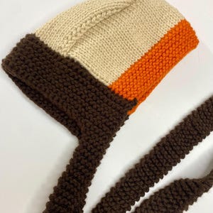 May include: A hand-knitted bonnet featuring a color-blocked design with a cream-colored crown, a vibrant orange middle section, and a dark brown border and long chin ties. The bonnet is constructed with a textured garter stitch pattern throughout.