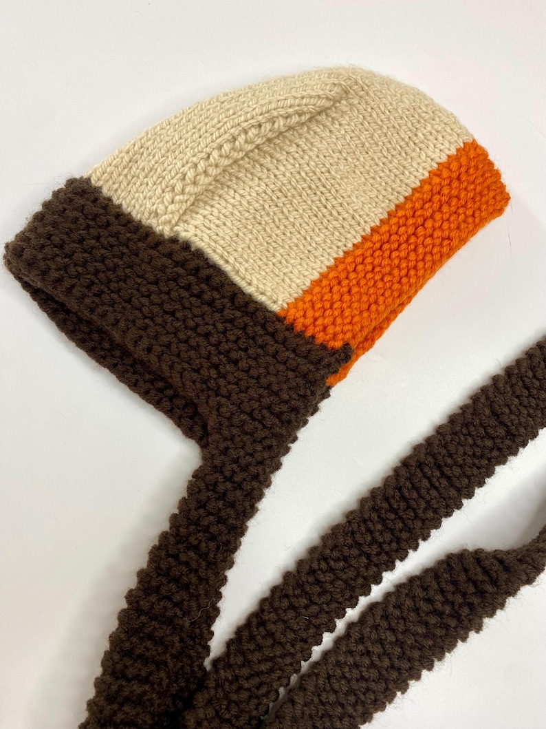 May include: A hand-knitted bonnet featuring a color-blocked design with a cream-colored crown, a vibrant orange middle section, and a dark brown border and long chin ties. The bonnet is constructed with a textured garter stitch pattern throughout.