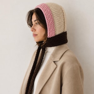 May include: A knit bonnet featuring a color-blocked design with a cream-colored crown, a pink border around the face, and a dark brown neck section with long, dangling knit ties. The bonnet is worn over a white ribbed top and a tan wool coat.