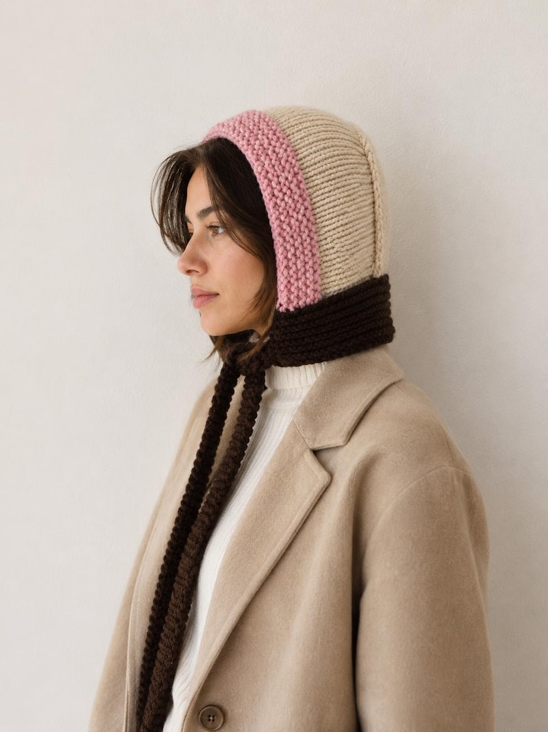 May include: A knit bonnet featuring a color-blocked design with a cream-colored crown, a pink border around the face, and a dark brown neck section with long, dangling knit ties. The bonnet is worn over a white ribbed top and a tan wool coat.