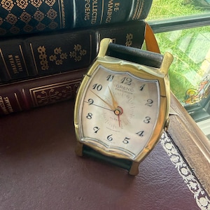 May include: A vintage brass alarm clock with a cream-colored face, black leather handle, and the text "GRENE PRECISION." The clock features a classic design with a gold-tone frame and black accents, resting on a dark surface near books.