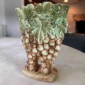 May include: A ceramic vase featuring a three-dimensional relief design of green grape leaves at the top and clusters of brown grapes forming the body. The piece has a glossy finish and stands on a rectangular base with a light tan color.