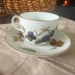 Puede incluir: Juego de taza y platillo blancos con borde dorado. La taza y el platillo están decorados con un diseño botánico con ciruelas moradas, hojas verdes y ramas marrones. El juego está sobre un mantel beige.
