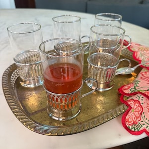 May include: A set of six clear glass cups, some filled with a red liquid, presented on a gold-toned metal tray. Each cup is held in a decorative silver-colored metal holder with a handle. A floral patterned cloth is on the side.