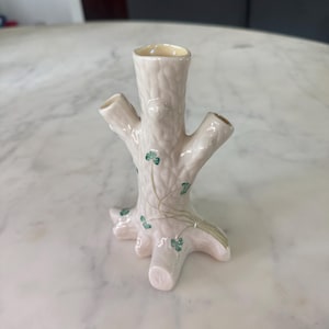 Pode incluir: Um vaso de cerâmica cor de creme em forma de tronco de árvore com três aberturas no topo. A superfície apresenta um padrão de casca texturizada e é decorada com pequenos trevos verdes pintados à mão e caules finos castanhos claros.