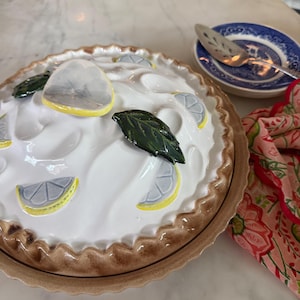 May include: A ceramic pie plate with a white, textured top, decorated with lemon slices and green leaves. The pie plate has a brown crust and is next to a blue and white plate with a pie server and a floral napkin.
