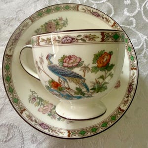 May include: Cream-colored teacup and saucer set with a detailed bird and floral design. The cup features a blue and teal bird with pink and orange flowers. The saucer and cup rims have a brown border with a green and brown geometric pattern.
