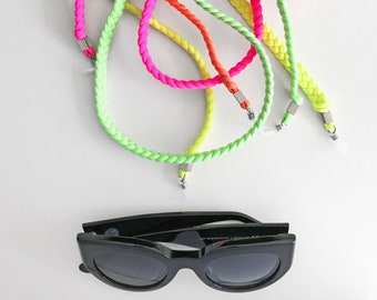 May include: A set of five neon colored braided eyeglass straps. The straps are in shades of green, yellow, orange, and pink. The straps are made of a woven material and have metal clasps at the ends. A pair of black sunglasses is also shown in the image.
