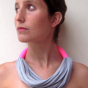 以下が含まれることがあります： 明るいピンク色の襟が付いたグレーの生地のネックレス。 ネックレスは、柔らかい素材の複数のストランドでできており、重ね着効果を生み出しています。 画像の女性は、濃い青色のストラップレスドレスを着ています。