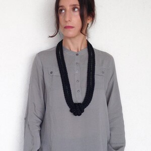 May include: A woman wearing a gray long-sleeved shirt with a black knotted necklace. The shirt has a button-up collar and two pockets.