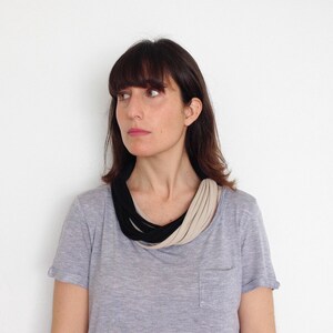 May include: A woman with dark brown hair is wearing a gray t-shirt with a black and beige fabric necklace.