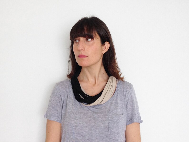 May include: A woman with dark brown hair is wearing a gray t-shirt with a black and beige fabric necklace.