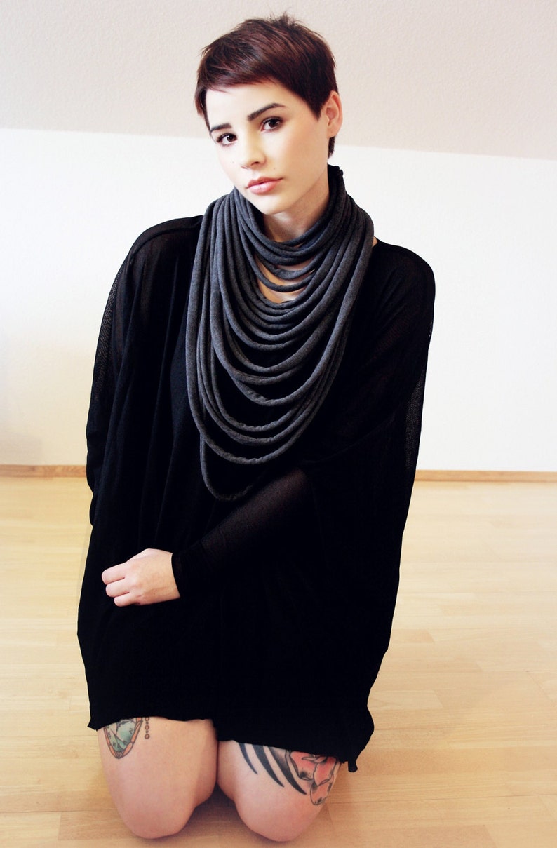May include: A woman with short, dark brown hair is wearing a black long-sleeved shirt with a grey infinity scarf. She is kneeling on a wooden floor and has tattoos on both legs.