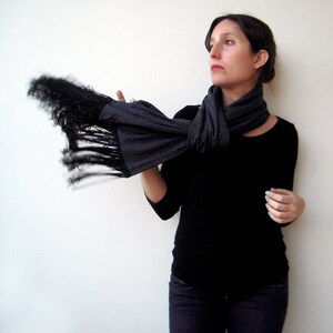 May include: A woman wearing a black long-sleeved shirt and dark wash jeans is wearing a gray scarf with black fringe. The scarf is tied around her neck and the fringe is flowing in the air.