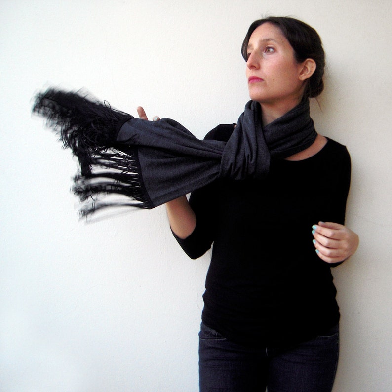 May include: A woman wearing a black long-sleeved shirt and dark wash jeans is wearing a gray scarf with black fringe. The scarf is tied around her neck and the fringe is flowing in the air.