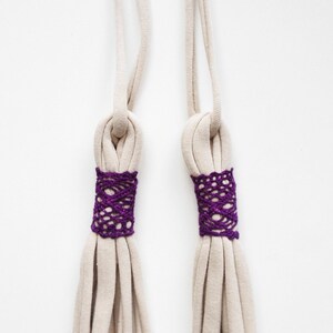 May include: Two beige fabric cords with purple crocheted trim at the ends. The cords are tied together in a knot at the top.