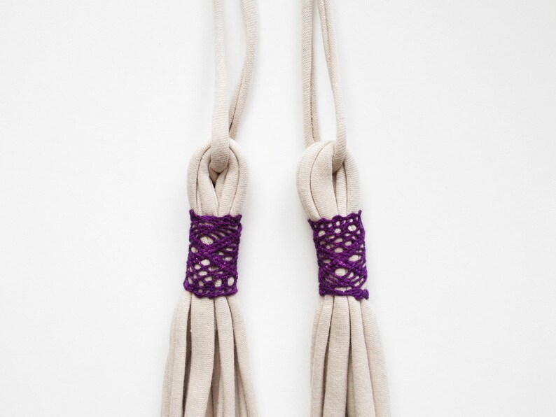May include: Two beige fabric cords with purple crocheted trim at the ends. The cords are tied together in a knot at the top.