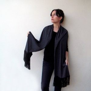May include: A charcoal gray shawl with a long fringe. The shawl is draped around a person's shoulders and is worn with a black top and dark jeans.