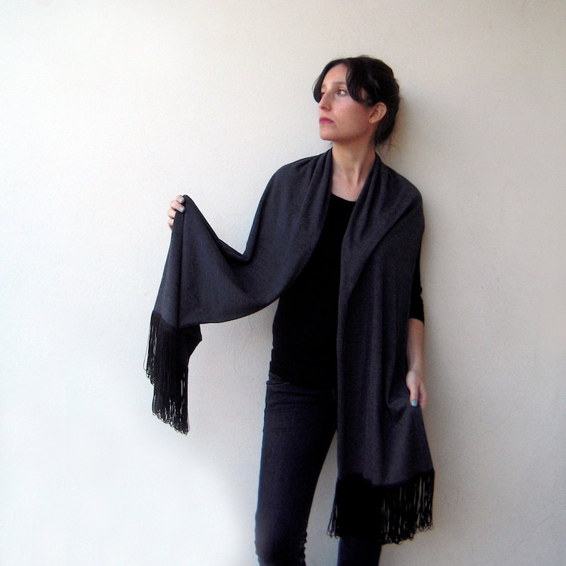 May include: A charcoal gray shawl with a long fringe. The shawl is draped around a person's shoulders and is worn with a black top and dark jeans.