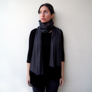 May include: A woman wearing a black shirt and a gray scarf with black fringe. The scarf is folded and worn around the neck.