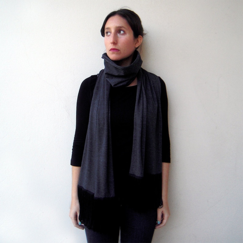 May include: A woman wearing a black shirt and a gray scarf with black fringe. The scarf is folded and worn around the neck.