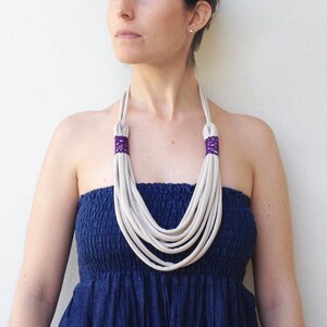 May include: A white multi-strand necklace with purple accents. The necklace is made of fabric and is worn by a person with a blue strapless dress.