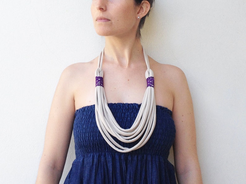May include: A white multi-strand necklace with purple accents. The necklace is made of fabric and is worn by a person with a blue strapless dress.