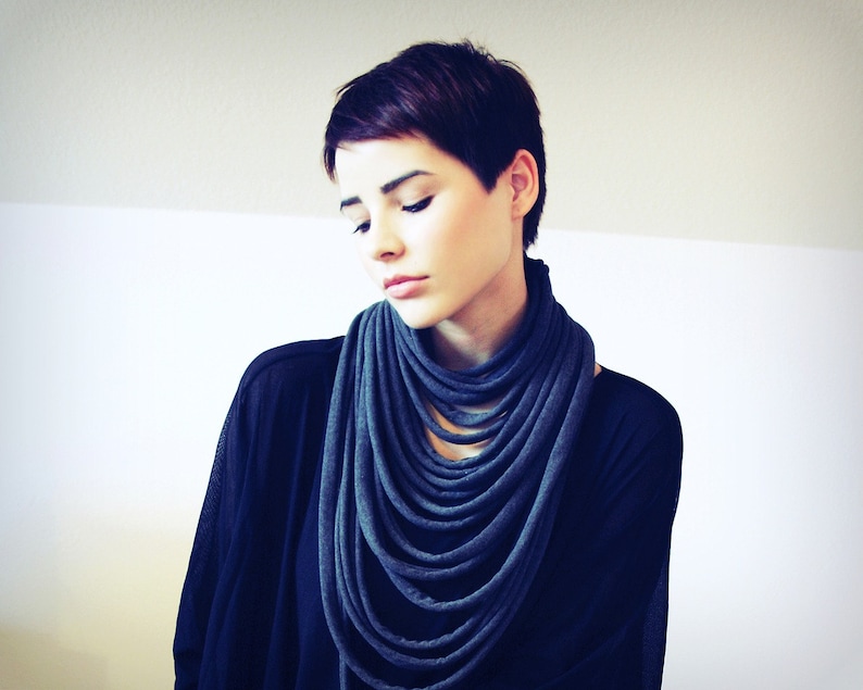 May include: A woman with short dark hair is wearing a dark blue infinity scarf. The scarf is made of a soft, stretchy fabric and is worn around the neck.