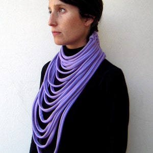 May include: A woman wearing a black turtleneck sweater with a purple fabric necklace. The necklace is made of multiple layers of fabric, creating a thick and voluminous look.