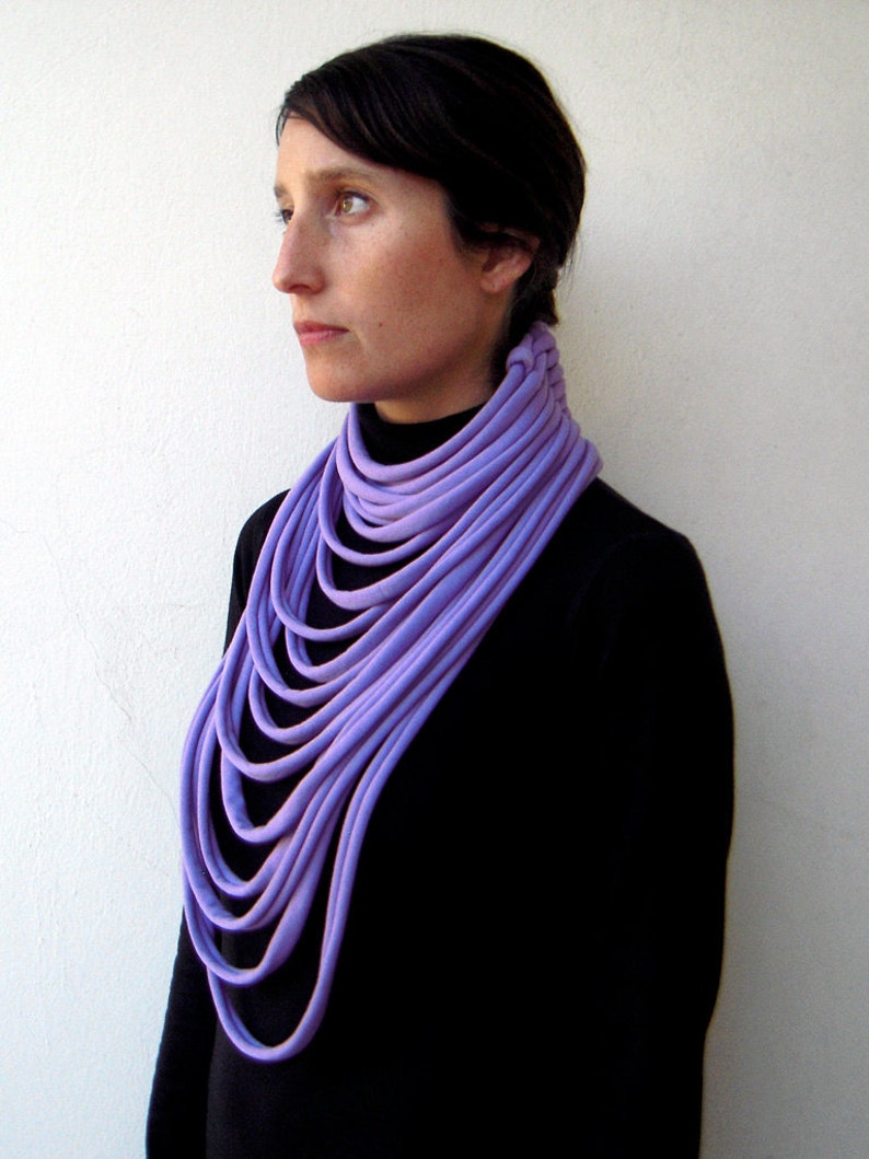 May include: A woman wearing a black turtleneck sweater with a purple fabric necklace. The necklace is made of multiple layers of fabric, creating a thick and voluminous look.