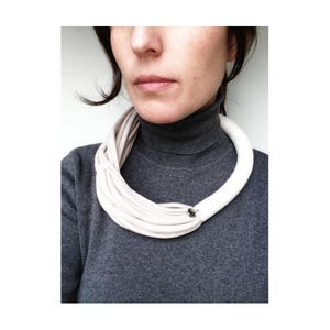May include: A white fabric necklace with a simple metal clasp. The necklace is made of multiple layers of fabric, creating a thick, chunky look. The necklace is worn by a person wearing a gray turtleneck sweater.