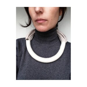 May include: A white fabric choker necklace with a gray fabric lining. The necklace is worn by a person with a gray turtleneck sweater.