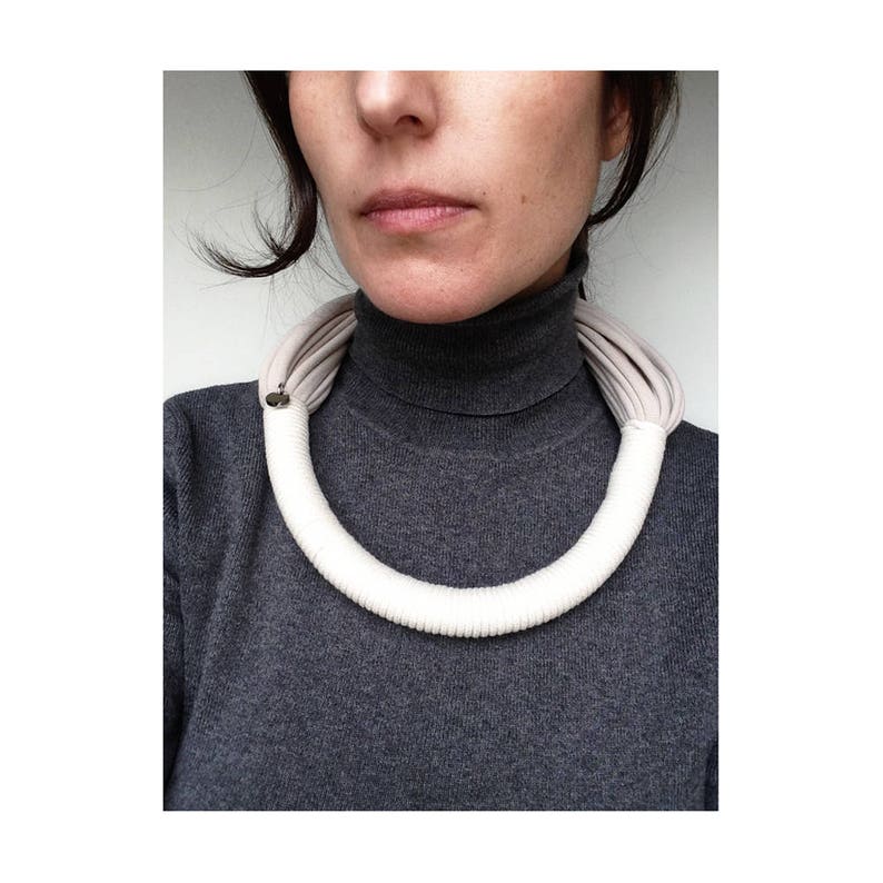 May include: A white fabric choker necklace with a gray fabric lining. The necklace is worn by a person with a gray turtleneck sweater.
