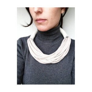 May include: A white fabric necklace with a simple silver clasp. The necklace is made of multiple layers of fabric, creating a soft and drapey look.